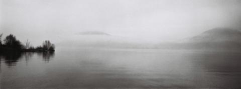 Visuel de l'exposition "L'ombre des roseaux" sur laquelle on observe une photo en noir et blanc d'une étendue d'eau, avec des collines en arrière-plan.